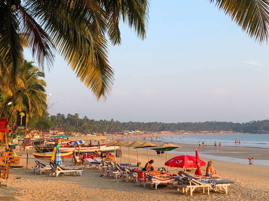 licensed-image: Palolem Beach, Goa, India - November, 28 2018: Stock photo of beach hut shack under tropical coconut palm tree at Palolem Beach, Goa, India. Sunbathers on holiday vacation sun loungers, holidaymakers swimming in sea, kayaks, canoes, fishing boats and parasol umbrellas.