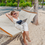 Workation: Woman in sunglasses relaxing on a hammock with a laptop on a tropical beach - concept of remote work or digital nomad lifestyle