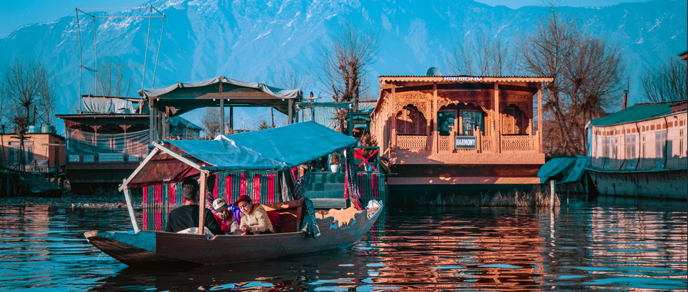 Top 10 Places to Visit in North India: A traditional wooden boat on a serene body of water. A rower is inside the boat, which is adorned with colorful fabrics and patterns. In the background, there’s an intricately designed wooden building alongside other structures. Snow-capped mountains rise majestically in the distance under a clear blue sky. The scene captures both cultural and natural elements, suggesting a place rich in tradition and natural beauty. 🌊🏔️