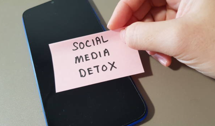 Hand holding a pink sticky note with ‘SOCIAL MEDIA DETOX’ written on it, covering the screen of a smartphone.